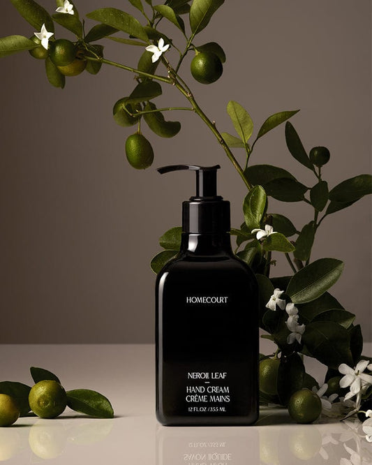 Black Homecourt Neroli Hand Cream bottle with neroli branches and fruit around it on a reflective surface.