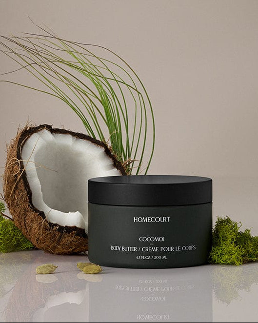 Black jar of Homecourt Indulgent Body Butter with a coconut and greenery on a neutral background.